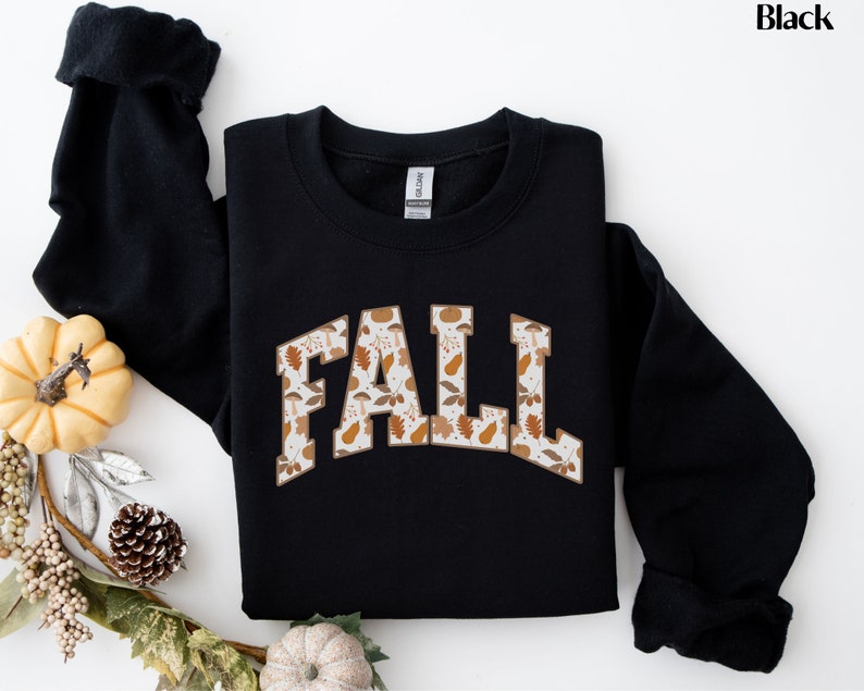 K&ouml;nnte beinhalten: Schwarzer Pullover mit einem wei&szlig;en "FALL"-Motiv mit einem Muster aus Herbstbl&auml;ttern und K&uuml;rbissen.