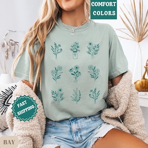 May include: A light green t-shirt with a floral design. The design features nine different floral arrangements, each with a unique combination of flowers and leaves. The flowers are outlined in a dark teal color.