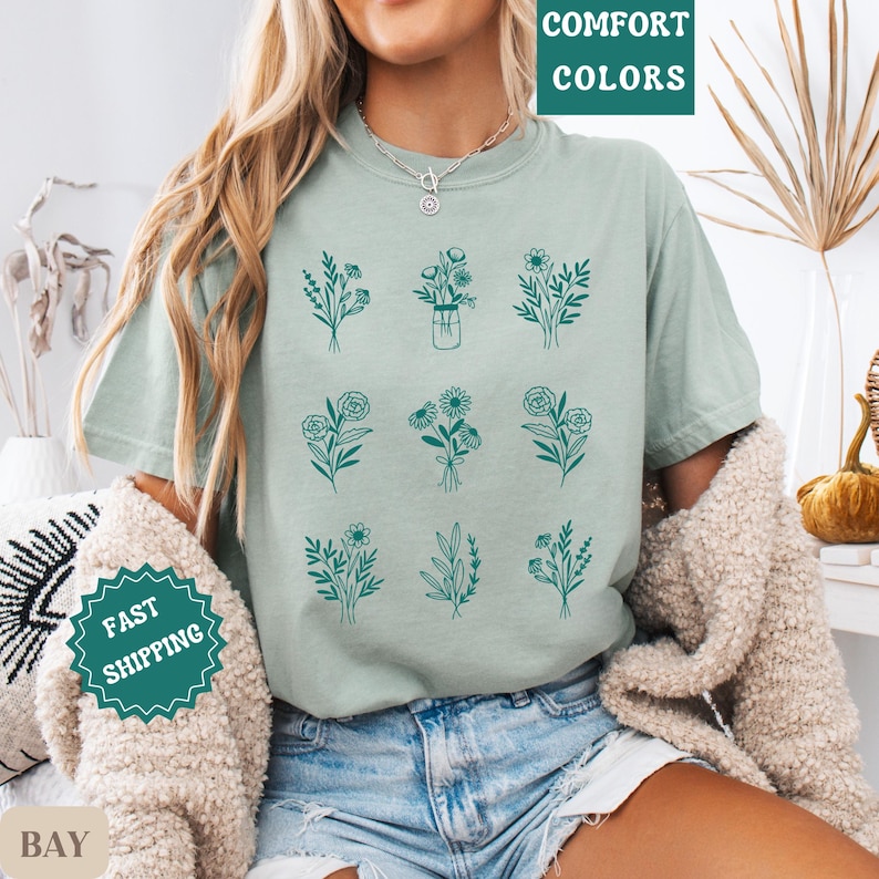 May include: A light green t-shirt with a floral design. The design features nine different floral arrangements, each with a unique combination of flowers and leaves. The flowers are outlined in a dark teal color.