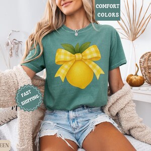 Puede incluir: Una camiseta verde claro con un lim&oacute;n amarillo y un lazo de cuadros amarillos y blancos. El texto "COMFORT COLORS" est&aacute; en la esquina superior derecha de la imagen.