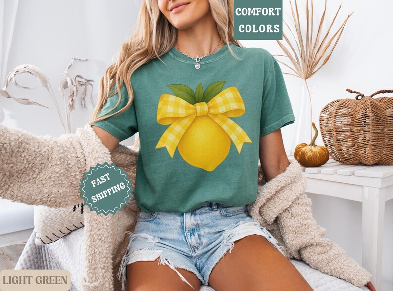 Puede incluir: Una camiseta verde claro con un lim&oacute;n amarillo y un lazo de cuadros amarillos y blancos. El texto "COMFORT COLORS" est&aacute; en la esquina superior derecha de la imagen.