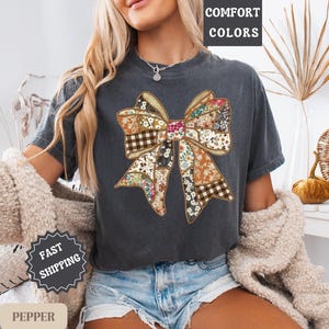 May include: Dark gray t-shirt featuring a large bow design with a patchwork pattern of floral, plaid, and solid fabrics. The words "COMFORT COLORS" are printed at the top. The shirt is worn with denim shorts.