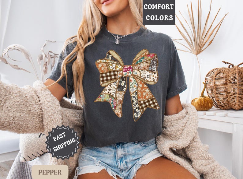 May include: Dark gray t-shirt featuring a large bow design with a patchwork pattern of floral, plaid, and solid fabrics. The words "COMFORT COLORS" are printed at the top. The shirt is worn with denim shorts.