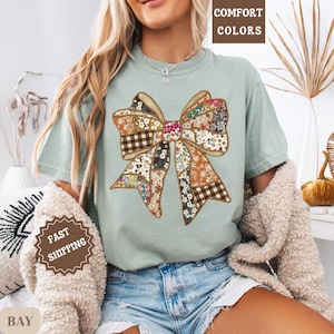 May include: A light green t-shirt featuring a large bow design. The bow is made of various fabric patterns, including floral, plaid, and solid colors. The words "COMFORT COLORS" are printed at the top. The words "FAST SHIPPING" are printed on a sticker.