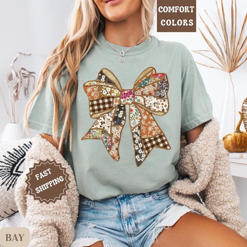 May include: A light green t-shirt featuring a large bow design. The bow is made of various fabric patterns, including floral, plaid, and solid colors. The words "COMFORT COLORS" are printed at the top. The words "FAST SHIPPING" are printed on a sticker.