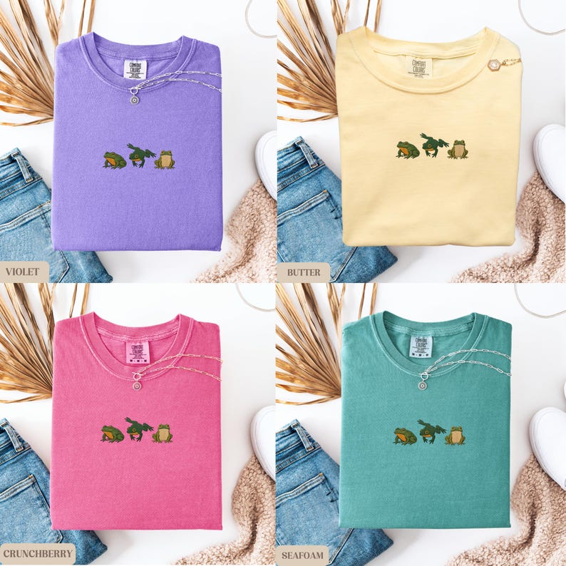 May include: Four t-shirts in violet, butter, crunchberry, and seafoam colors, each with an embroidered frog design. The shirts are displayed with jeans, dried plants, and white sneakers.