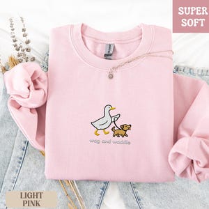 May include: Light pink sweatshirt with a crew neck. The front features an embroidered design of a duck walking a dog on a leash, with the words "wag and waddle" below. The words "Super Soft" and "Light Pink" are also visible.