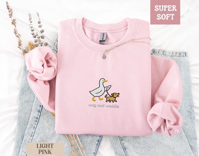 May include: Light pink sweatshirt with a crew neck. The front features an embroidered design of a duck walking a dog on a leash, with the words "wag and waddle" below. The words "Super Soft" and "Light Pink" are also visible.