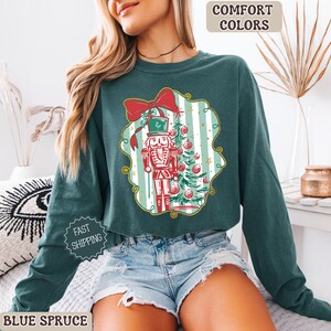 May include: A teal long-sleeve sweatshirt with a Christmas-themed graphic. The graphic features a nutcracker, Christmas tree, and a red bow. The text "COMFORT COLORS" is at the top, and "BLUE SPRUCE" is at the bottom.