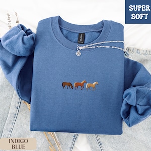 K&ouml;nnte beinhalten: Indigoblaues Sweatshirt mit gestickten Pferden. Das Oberteil mit Rundhalsausschnitt tr&auml;gt die Worte "SUPER SOFT" in einer blauen Box. Eine silberne Halskette mit Anh&auml;nger befindet sich auf dem Shirt. Die &Auml;rmel sind gefaltet. Das Sweatshirt liegt auf einer Jeans.