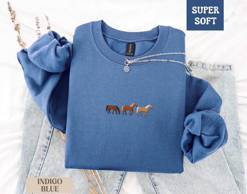 K&ouml;nnte beinhalten: Indigoblaues Sweatshirt mit gestickten Pferden. Das Oberteil mit Rundhalsausschnitt tr&auml;gt die Worte "SUPER SOFT" in einer blauen Box. Eine silberne Halskette mit Anh&auml;nger befindet sich auf dem Shirt. Die &Auml;rmel sind gefaltet. Das Sweatshirt liegt auf einer Jeans.