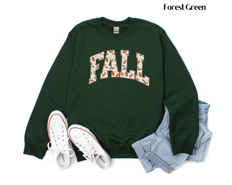 May include: Forest green sweatshirt with the word "FALL" in a white, arched design filled with fall leaves. White sneakers and blue jeans are next to the sweatshirt. The sweatshirt is a crew neck style.