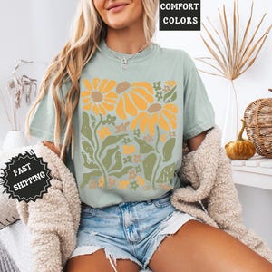 May include: A sage green short-sleeve t-shirt featuring a graphic print of large yellow sunflowers and green foliage. The shirt has a crew neckline and is worn with light blue denim shorts and a textured beige cardigan draped over the shoulders.