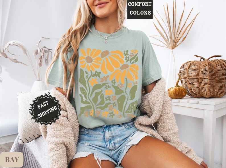 May include: A sage green short-sleeve t-shirt featuring a graphic print of large yellow sunflowers and green foliage. The shirt has a crew neckline and is worn with light blue denim shorts and a textured beige cardigan draped over the shoulders.