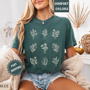 May include: A teal green t-shirt with a graphic of nine different floral arrangements in white. The text "COMFORT COLORS" is printed on the top left corner of the shirt.