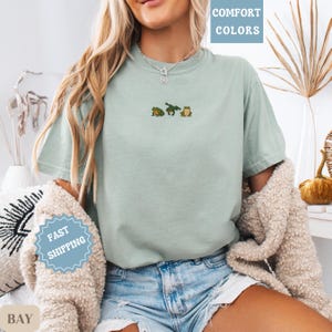 May include: A light green t-shirt with three embroidered frogs. The shirt has the words "COMFORT COLORS" printed in a box in the upper right corner. The shirt is paired with denim shorts.