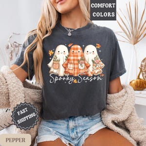 May include: Dark grey t-shirt with a design of three cartoon ghosts, each with floral and plaid details, holding baskets. The words "Spooky Season" are written below. The t-shirt has a vintage, worn appearance.