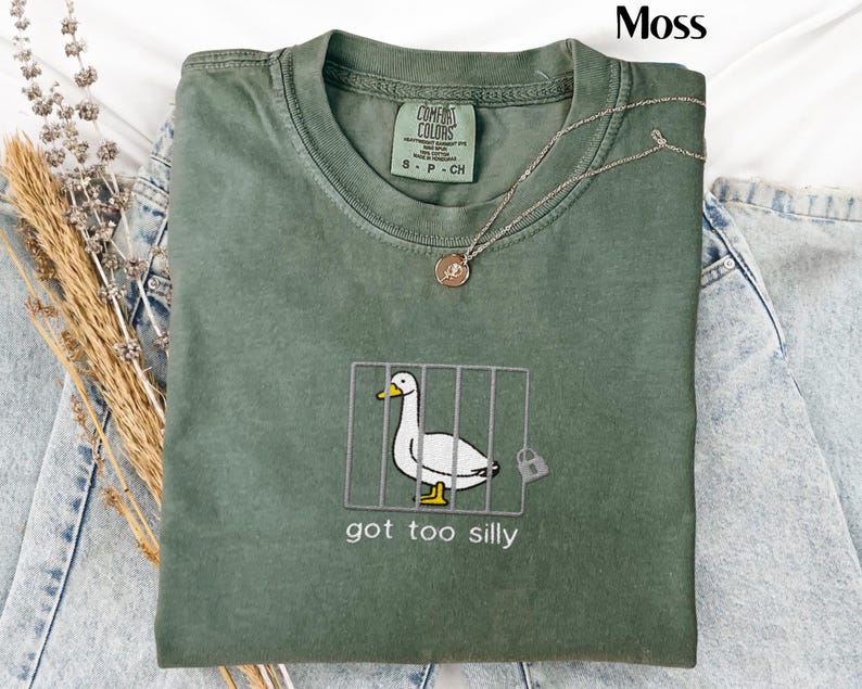 May include: A moss green t-shirt with a graphic of a white duck in a cage, the text "got too silly" is below the graphic. The shirt is folded and has a silver necklace on top. The shirt is made by Comfort Colors.