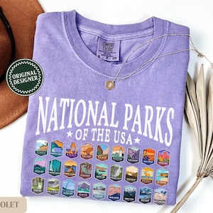 以下が含まれることがあります： 紫色のTシャツに「National Parks of the USA」という白い文字が書かれています。Tシャツには、さまざまな国立公園を表すカラフルなパッチがグリッド状に並んでいます。