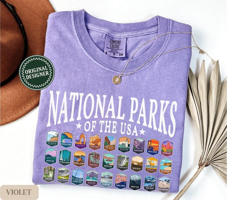 以下が含まれることがあります： 紫色のTシャツに「National Parks of the USA」という白い文字が書かれています。Tシャツには、さまざまな国立公園を表すカラフルなパッチがグリッド状に並んでいます。