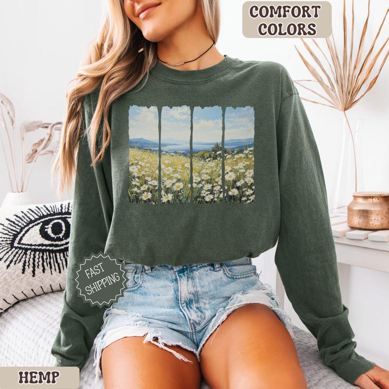 May include: An olive green sweatshirt featuring a scenic print of a field of daisies under a blue sky. The graphic is framed by vertical lines, creating a window effect. The words "COMFORT COLORS" and "HEMP" are printed on the image.
