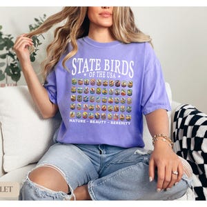 May include: A woman wearing a purple t-shirt with the text "State Birds of the USA" and a graphic of birds. The shirt also has the text "Nature - Beauty - Serenity".