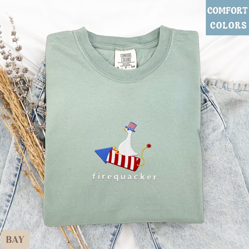 K&ouml;nnte beinhalten: Hellgr&uuml;nes T-Shirt mit Rundhalsausschnitt und kurzen &Auml;rmeln. Das Shirt hat ein gesticktes Design einer Gans mit patriotischem Hut, die auf einer Rakete reitet, mit dem Wort "firequacker" darunter. Das T-Shirt liegt auf einer Jeans.