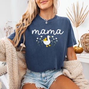 May include: A navy blue t-shirt with the word "mama" printed in white. A white duck with a yellow chick on its back is printed below the text. The duck is surrounded by white flowers.