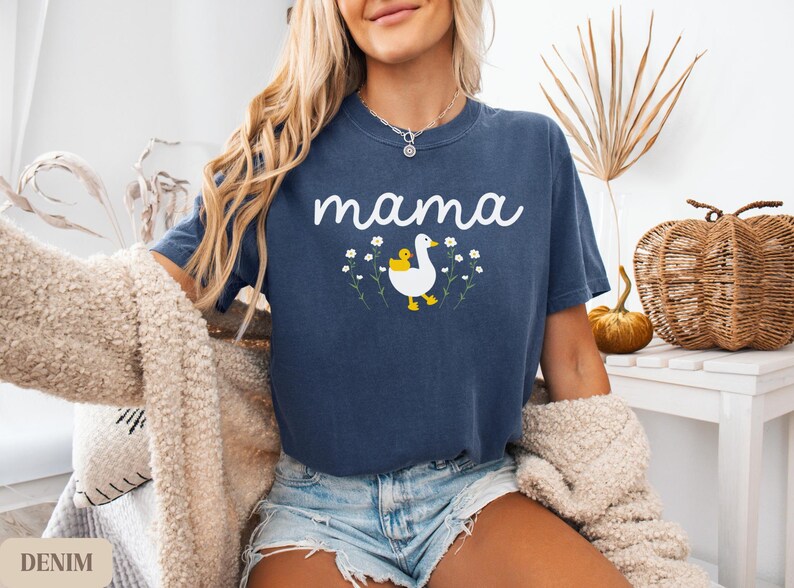 May include: A navy blue t-shirt with the word "mama" printed in white. A white duck with a yellow chick on its back is printed below the text. The duck is surrounded by white flowers.