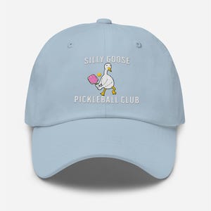 May include: Light blue baseball cap with white embroidery that reads "Silly Goose Pickleball Club". The embroidery also includes a cartoon goose holding a pink pickleball paddle.