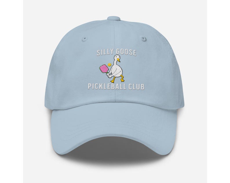May include: Light blue baseball cap with white embroidery that reads "Silly Goose Pickleball Club". The embroidery also includes a cartoon goose holding a pink pickleball paddle.