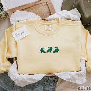 May include: A pale yellow sweatshirt with three green crocodile designs. The sweatshirt is presented in a wooden tray with white tissue paper and a small card that says "For you". A green hat is visible in the background.
