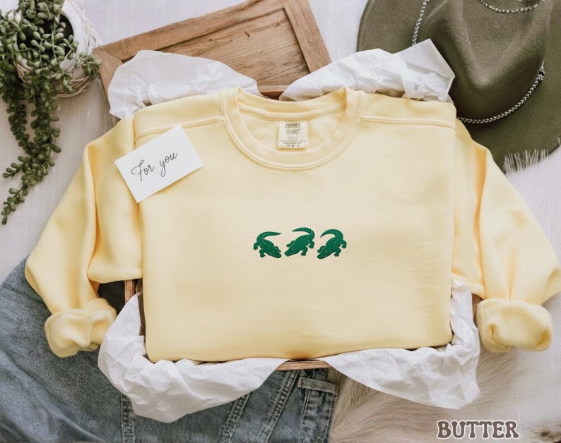 May include: A pale yellow sweatshirt with three green crocodile designs. The sweatshirt is presented in a wooden tray with white tissue paper and a small card that says "For you". A green hat is visible in the background.