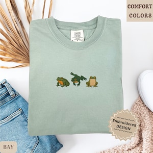 May include: A light green Comfort Colors t-shirt with an embroidered design of three frogs. The frogs are in shades of green and brown. The shirt has a crew neck and short sleeves. The text "COMFORT COLORS" is visible.