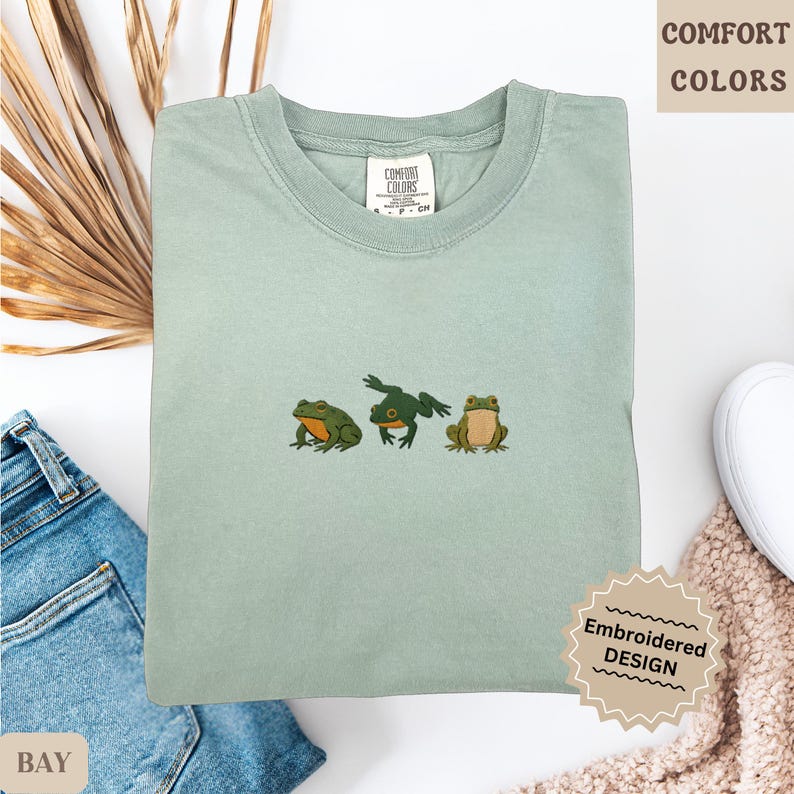 May include: A light green Comfort Colors t-shirt with an embroidered design of three frogs. The frogs are in shades of green and brown. The shirt has a crew neck and short sleeves. The text "COMFORT COLORS" is visible.