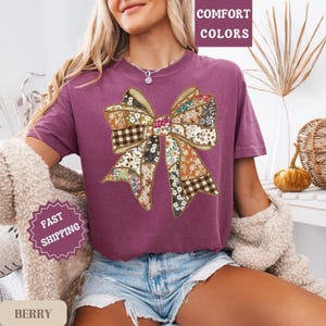 May include: Berry-colored t-shirt featuring a patchwork bow design. The bow incorporates floral and plaid patterns in various colors. The shirt has the words "Comfort Colors" and "Fast Shipping" printed on it.