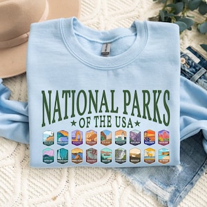 May include: Light blue sweatshirt with the text "National Parks of the USA" in green lettering. The sweatshirt features a row of colorful patches depicting various national parks.
