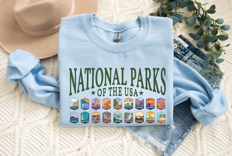 May include: Light blue sweatshirt with the text "National Parks of the USA" in green lettering. The sweatshirt features a row of colorful patches depicting various national parks.