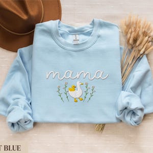 May include: Light blue sweatshirt with the word "mama" embroidered in white. A white duck with a yellow duckling is embroidered below the word. The duck and duckling are surrounded by white flowers.