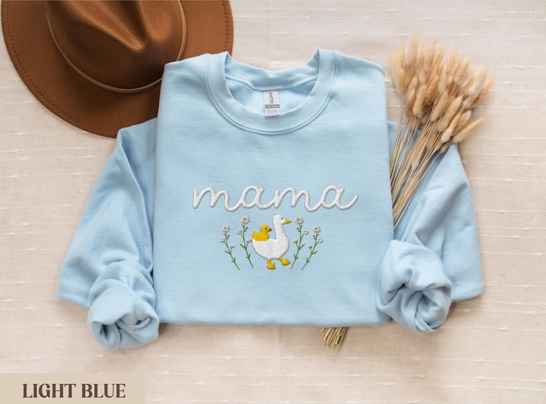May include: Light blue sweatshirt with the word "mama" embroidered in white. A white duck with a yellow duckling is embroidered below the word. The duck and duckling are surrounded by white flowers.