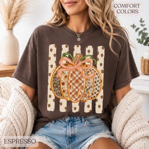 May include: A dark brown short-sleeve t-shirt featuring a graphic of a pumpkin with a pink bow. The pumpkin has sections with leopard print, plaid, and floral patterns. Behind the pumpkin are vertical stripes with polka dots. Text reads COMFORT COLORS.