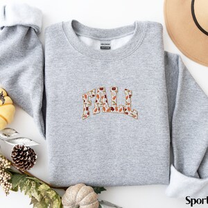 May include: A gray sweatshirt with a "FALL" graphic made of autumn leaves. The sweatshirt is a crew neck style and has a relaxed fit.
