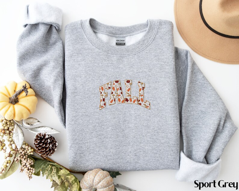 May include: A gray sweatshirt with a "FALL" graphic made of autumn leaves. The sweatshirt is a crew neck style and has a relaxed fit.