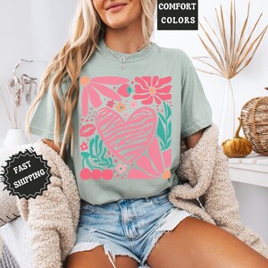May include: Sage green t-shirt with a pink and white floral heart design. The graphic includes flowers, a strawberry, cherries, and a kiss. The words "COMFORT COLORS" are in a black box at the top.
