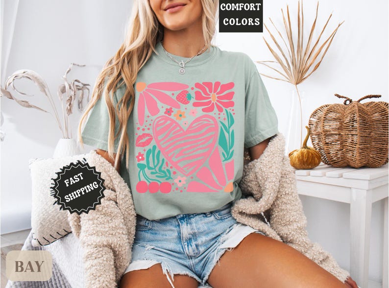 May include: Sage green t-shirt with a pink and white floral heart design. The graphic includes flowers, a strawberry, cherries, and a kiss. The words "COMFORT COLORS" are in a black box at the top.