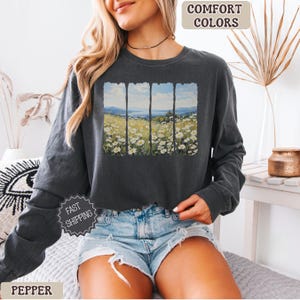 May include: Dark gray long-sleeve sweatshirt with a landscape print. The print features a field of white flowers, a blue sky, and mountains. The words "Comfort Colors" are printed at the top. The word "Pepper" is printed at the bottom.
