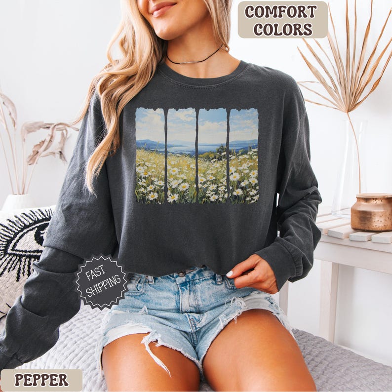 May include: Dark gray long-sleeve sweatshirt with a landscape print. The print features a field of white flowers, a blue sky, and mountains. The words "Comfort Colors" are printed at the top. The word "Pepper" is printed at the bottom.