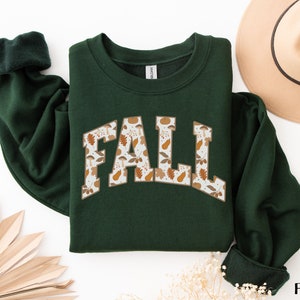 K&ouml;nnte beinhalten: Ein dunkelgr&uuml;ner Pullover mit dem Wort "FALL" auf der Vorderseite in wei&szlig;er Umrandung. Die Buchstaben sind mit einem Muster aus braunen Bl&auml;ttern, Eicheln und K&uuml;rbissen gef&uuml;llt.