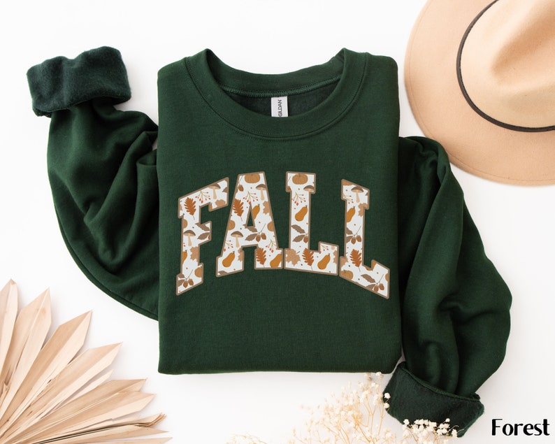 K&ouml;nnte beinhalten: Ein dunkelgr&uuml;ner Pullover mit dem Wort "FALL" auf der Vorderseite in wei&szlig;er Umrandung. Die Buchstaben sind mit einem Muster aus braunen Bl&auml;ttern, Eicheln und K&uuml;rbissen gef&uuml;llt.