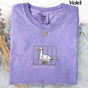 May include: A violet Comfort Colors t-shirt with a graphic of a duck in a cage, a padlock, and the text "got too silly." A silver necklace with a pendant is on the shirt.